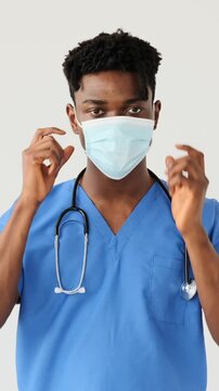 Vertical video of a young African American doctor in scrubs taking off his protective face mask, isolated on a white background.