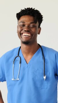 Vertical video of a cheerful African American male doctor in blue scrubs with a stethoscope smiling at the camera, isolated on a white background.