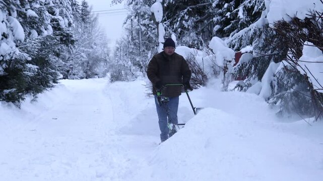 Man clearing deep snow with a snow blower on a winter residential road surrounded by snow-covered pine trees. Cold weather, heavy snowfall, winter maintenance and daily life in rural area