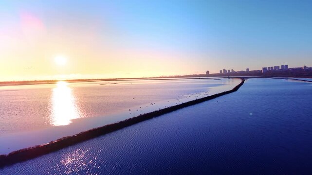 Geometric lines of salt pans and dikes stretching toward a distant city under a bright sun. Bright blue waters of lakes with birds. Picturesque sunset. Aerial view.