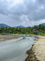 Serene fishing village scene on South Java coast under cloudy skies, shallow river mouth flowing into sandy beach, palm trees lining the shore, distant blue boats, and misty mountains. Rajegwesi Beach
