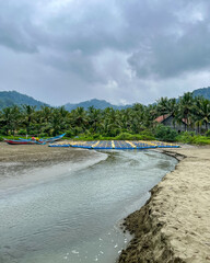 Serene fishing village scene on South Java coast under cloudy skies, shallow river mouth flowing into sandy beach, palm trees lining the shore, distant blue boats, and misty mountains. Rajegwesi Beach