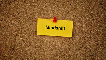 A yellow piece of paper with the word Mindshift on it pinned to a cork board. Close up.