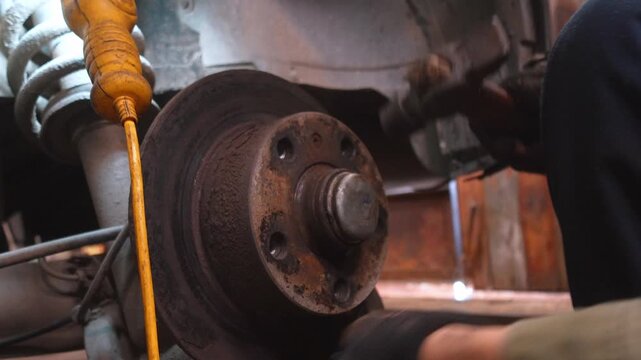 Professional auto mechanic wearing protective gloves using a hammer to remove an old rusty brake disc during car maintenance service in a repair shop, showing the vehicle suspension
