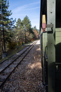 A scenic view from a vintage train window looking down parallel railroad tracks bordered by autumn leaves and tall pine trees under a clear blue sky. 