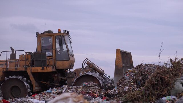 Heavy industrial machinery working at a large landfill site, with a special bulldozer compacting and managing piles of garbage and solid waste against a gray, overcast sky