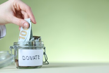 Woman putting dollar banknote into donation jar at white table against light green background,...