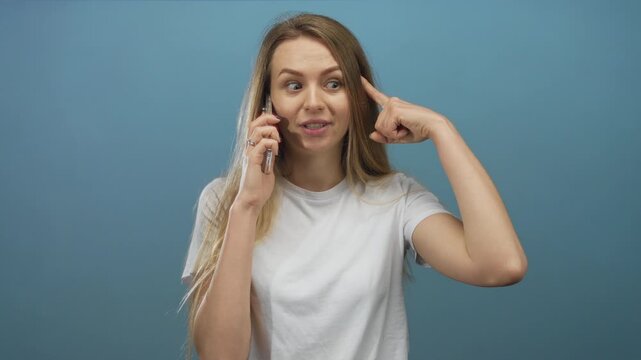 Woman talking on smartphone over blue background, expressing surprise and pointing at head while wearing casual white shirt.