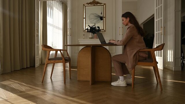 Young focused woman in a stylish suit sitting at a wooden table in a sunlit room, typing on a laptop and then checking her smartphone, working remotely in a modern elegant home office