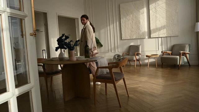 Elegant woman carrying a travel bag walking through a chic, sunlit apartment with modern furniture and herringbone parquet flooring, evoking a sense of travel, homecoming, or departure