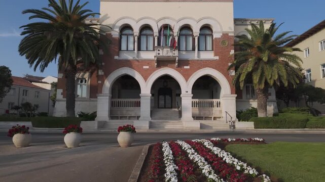 The beautiful Town Hall of Porec, Croatia on a sunny summer afternoon. Built in 1910, designed by an Italian architect. Romantic street scenery with huge old palm trees, and red and white flowers.