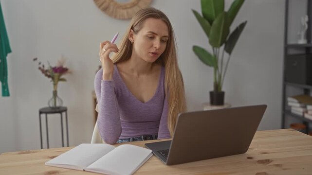 Young woman studying at home in a cozy living room, focused with a laptop and notebook, wearing a purple top, surrounded by books and plants.