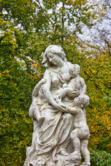 Obraz premium Bottom up perspective picture of a beautiful statue in the park, with a woman and her children. Marble sculpture over green and yellow foliage background.