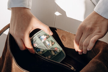 A hand holds a green, divided pill organizer filled with assorted tablets and capsules, set against a brown suede bag. The scene conveys health management, daily medications, and personal care.