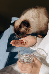 Cat Sniffs Pills From Hand While Holding Glass Of Water 