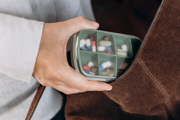 A hand holds a green, divided pill organizer filled with assorted tablets and capsules, set against a brown suede bag. The scene conveys health management, daily medications, and personal care.