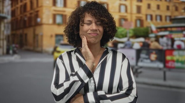 Woman pressing cheek with hand while wincing on a sunlit urban street with blurred building behind; discomfort.