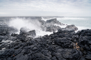 Waves crash on a stretch of coastline with black volcanic rocks and rough seas on the Reykjanes Peninsula in Iceland on a spring day.