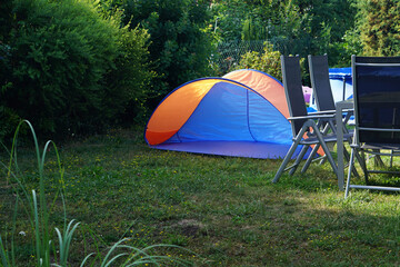 blue and orange pop up sun shelter tent set up on a green lawn in a backyard garden with patio chairs and bushes nearby for summer recreation.