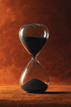 Clear glass hourglass with black sand in dystopian time concept on desaturated rust-orange background