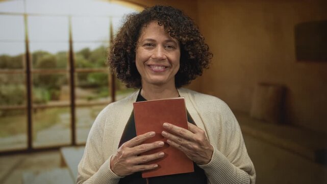 Woman tenderly holds journal to chest in rustic empty studio; comfort reflection serenity gratitude.
