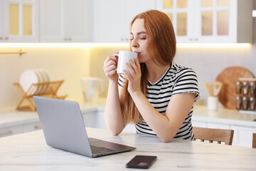 Woman having refreshing coffee while working on laptop at table in kitchen. Space for text