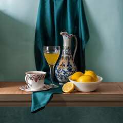 Still life with lemons, a wine glass, an elegant jug, and a cup of tea in yellow, white, blue, and green tones