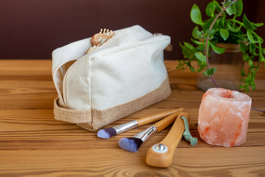 A natural linen beauty case on a wooden background with salt crystal candle holder a wooden roller for regenerating facial massage and bamboo makeup brushes. A zero-waste beauty concept