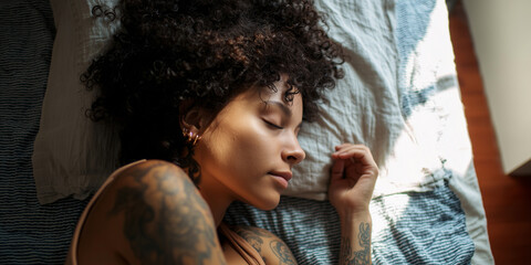 Beautiful young woman with curly hair and tattoos sleeping peacefully in bed during morning