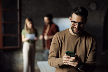 A professional man using his smartphone in the foreground while his colleagues work together in a blurred office background.