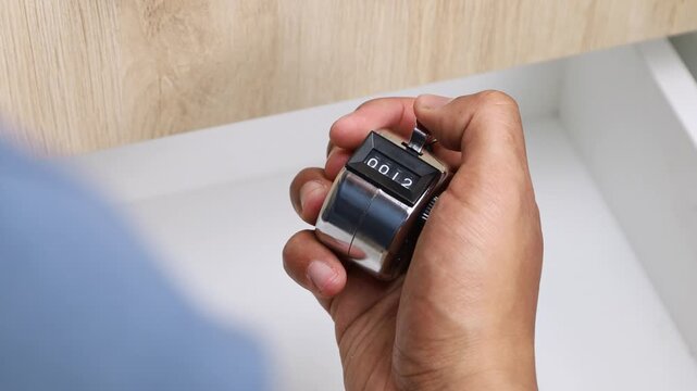 Close-up of a hand holding a mechanical tally counter used for counting events, people, or repetitions, showing a simple manual counting device on a neutral background.