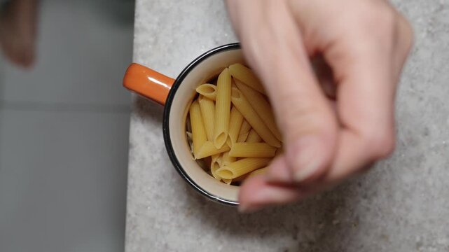 Top view of a hand placing dry penne pasta into a mug to measure portions before cooking. Simple kitchen scene representing meal preparation, portion control and home cooking
