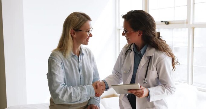 Middle-aged woman patient handshaking with female doctor gives consent to treatment plan, listening professional medical advice, reviewing together scan imaging results on tablet at visit at hospital