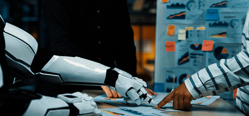 A futuristic scene depicts a collaboration between a human and a robot, analyzing charts and data in a modern office, highlighting innovation in technology and teamwork.Noogenesis