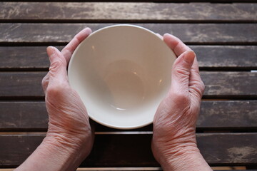 A old woman holding an empty bowl on a old wood dining table. Old hands holding an empty dish to eat. The price of food and the poverty among older adults. Cost of the living and elders.