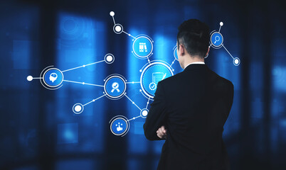 A business professional stands in a modern office, engaging with a digital display of technology icons and data connections, representing analysis and innovation. Tessel