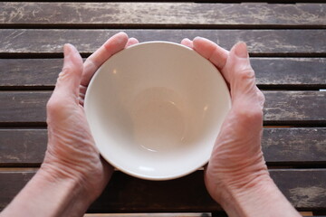 A old woman holding an empty bowl on a old wood dining table. Old hands holding an empty dish to eat. The price of food and the poverty among older adults. Cost of the living and elders.