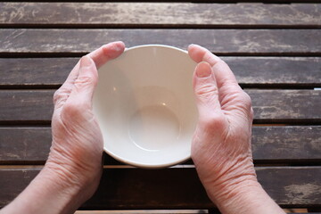 A old woman holding an empty bowl on a old wood dining table. Old hands holding an empty dish to eat. The price of food and the poverty among older adults. Cost of the living and elders.