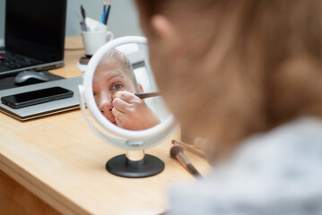 A middle aged woman applies makeup while looking into a circular vanity mirror on a wooden desk. Nearby laptop and phone suggest home office or personal studio