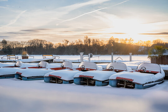 Bunte Tretboote liegen auf dem zugefrorenen Maschsee in Hannover bei Sonnenuntergang mit der Abendsonne im Winter mit Schnee und Eis 