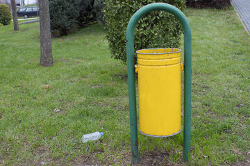 Crumpled plastic bottle on grass beside a trash bin in a public park. Symbol of pollution, negligence, and lack of environmental responsibility.