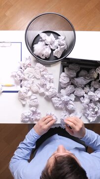 Vertical video frustrated person sitting at a white desk, surrounded by failed crumpled paper ideas and throwing another piece into a wastebasket, symbolizing stress and creative block in a workspace