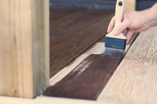 Brown oil for protection of wood by staining. Painting a larch for protecting and pests. Woodworker applying finish to terrace deck board. Repairman varnishing with brush wooden plank floor surface.