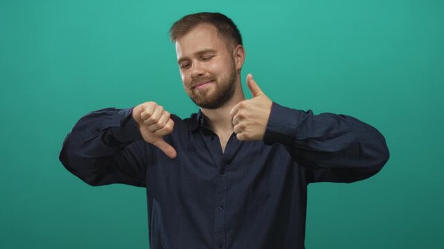 Young caucasian man showing thumbs up and thumbs down in studio against a teal backdrop with closed eyes and a subtle smile; ambivalence choice.