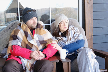 Couple relaxing on a patio wrapped on checkered blankets. Relaxed beautiful woman with a handsome man sitting on wooden cottage terrace. Winter resort. Happy loving couple enjoying leisure, embracing