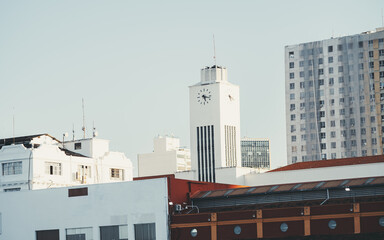 Urban skyline with modern white clock tower above low rooftops and a tall apartment block, clear pale blue sky, soft daylight, architectural cityscape with copy space