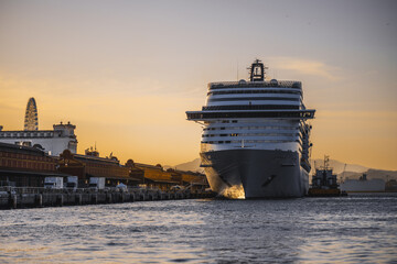 Huge cruise ship moored at harbor pier during golden sunset, front bow view near port terminal buildings, calm sea water, warm sky gradient and distant mountains