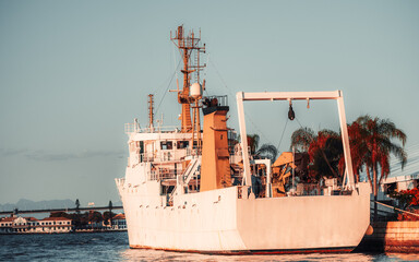 White industrial ship moored at harbor pier at sunset, stern view with crane gantry, mast antennas and bridge, calm water, palm trees and city coastline