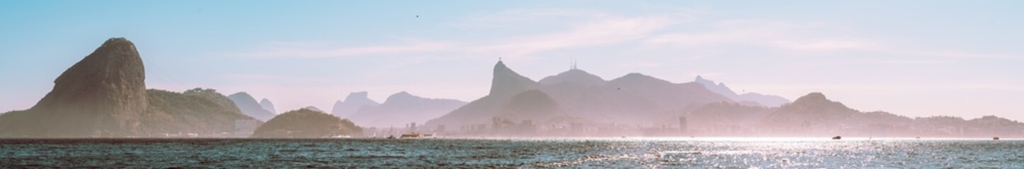 Obraz premium Panoramic Rio de Janeiro seascape with Sugarloaf Mountain and Corcovado silhouette, sparkling Guanabara Bay, pastel morning sky, haze and distant skyline, Brazil