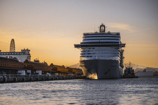 Huge cruise ship moored at harbor pier during golden sunset, front bow view near port terminal buildings, calm sea water, warm sky gradient and distant mountains - Powered by Adobe